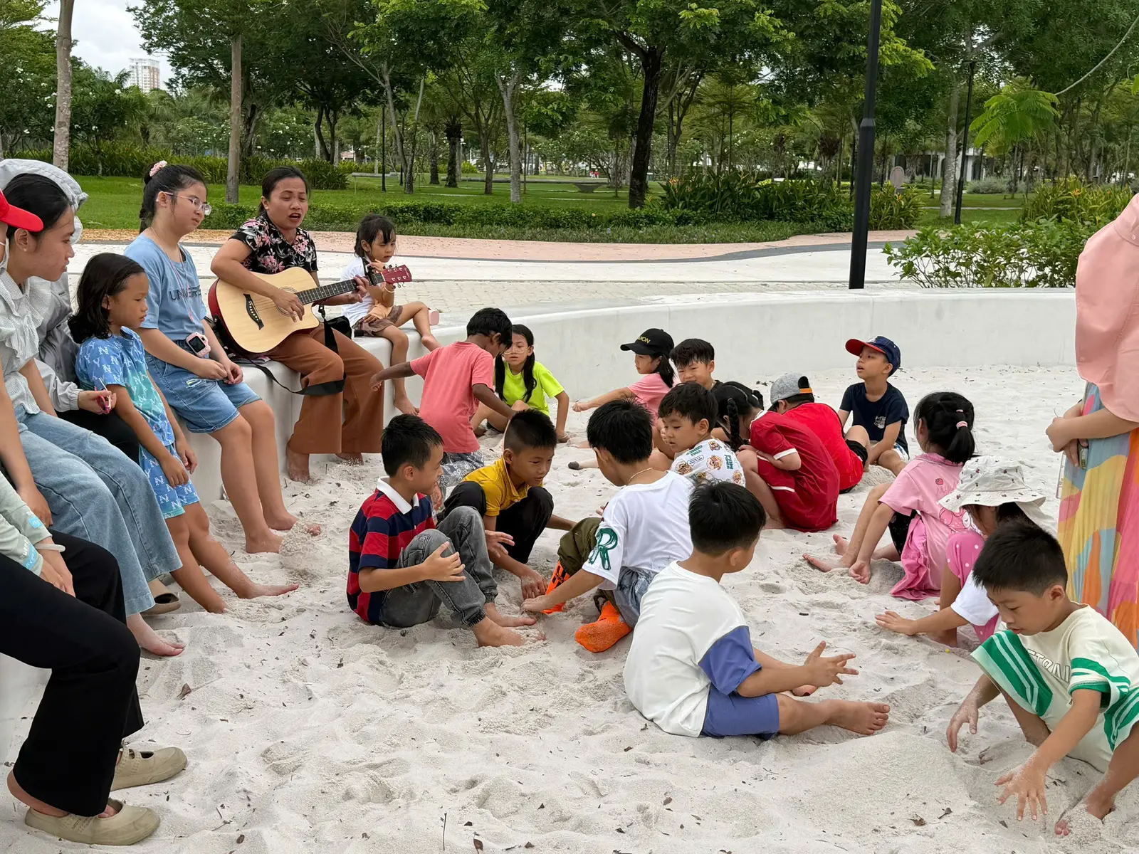Buổi học nghệ thuật ngoài trời tại một công viên, các bạn nhỏ đang vừa chơi vừa học, với tiếng đàn guitar rộn ràng của cô giáo.
